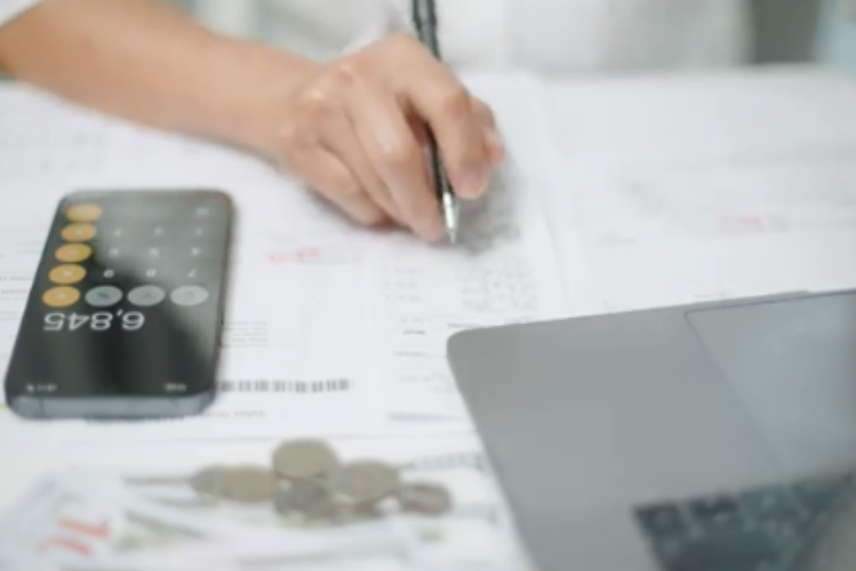 Hand writing on documents beside smartphone calculator and laptop with coins.